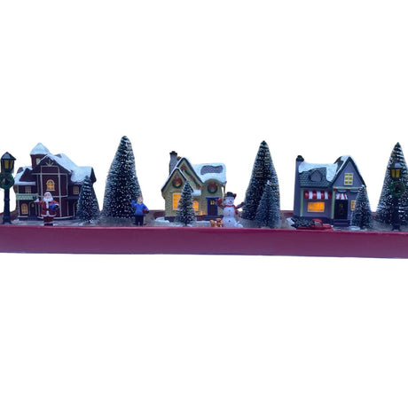 Set natalizio in miniatura con case illuminate, alberi innevati, Babbo Natale, pupazzo di neve e personaggi su confezione rossa.