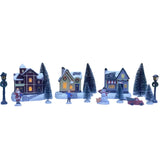 Set natalizio in miniatura con case illuminate, alberi innevati, Babbo Natale, pupazzo di neve e personaggi su sfondo bianco.