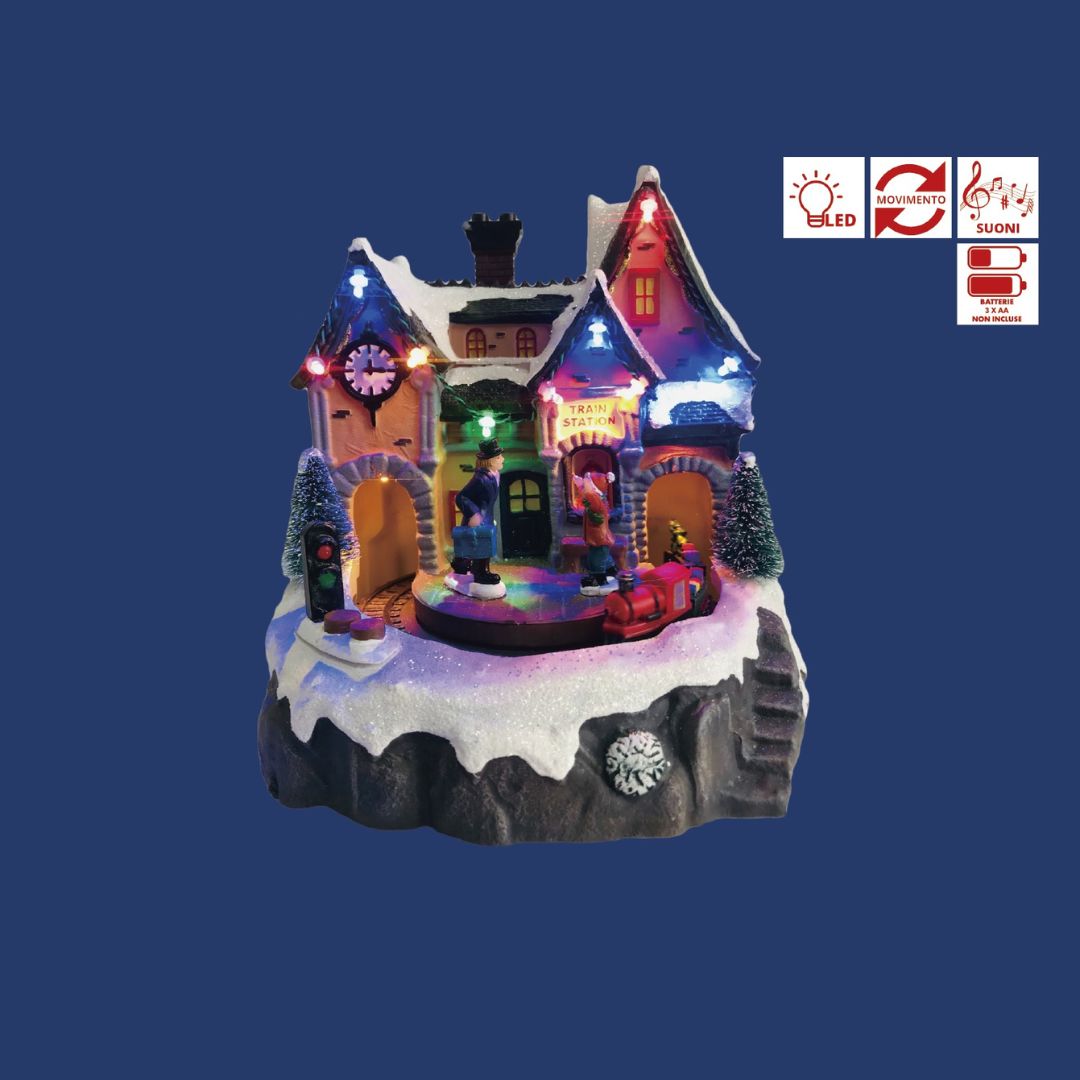 Stazione ferroviaria natalizia in miniatura con casa illuminata a LED, personaggi con valigie e trenino rosso sulla neve su sfondo blu.