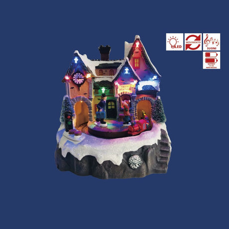 Stazione ferroviaria natalizia in miniatura con casa illuminata a LED, personaggi con valigie e trenino rosso sulla neve su sfondo blu.