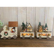 Trenino natalizio decorativo in legno con tre vagoni illuminati: locomotiva con pupazzo di neve, carrozza con casetta e personaggi natalizi e carrozza con renna e alberi di Natale.