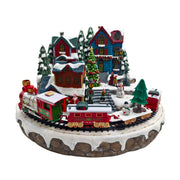 Villaggio natalizio in miniatura con case illuminate, albero decorato, pupazzi di neve e trenino rosso che gira intorno alla piazza innevata.