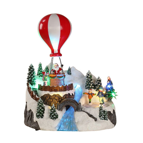 Villaggio natalizio in miniatura con Babbo Natale su mongolfiera, bambini che giocano sulla neve, alberi innevati e cascata illuminata.