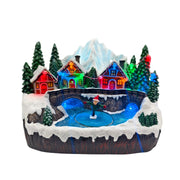 Villaggio natalizio in miniatura con case illuminate colorate, ponte in pietra, laghetto ghiacciato e pattinatore tra pini e montagne.