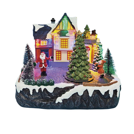 Villaggio natalizio in miniatura con casa illuminata, albero decorato, Babbo Natale e una bambina tra alberi innevati su sfondo bianco.