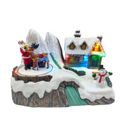 Villaggio natalizio in miniatura con Babbo Natale accanto a una renna, casa illuminata con LED multicolor, pupazzo di neve e paesaggio innevato.