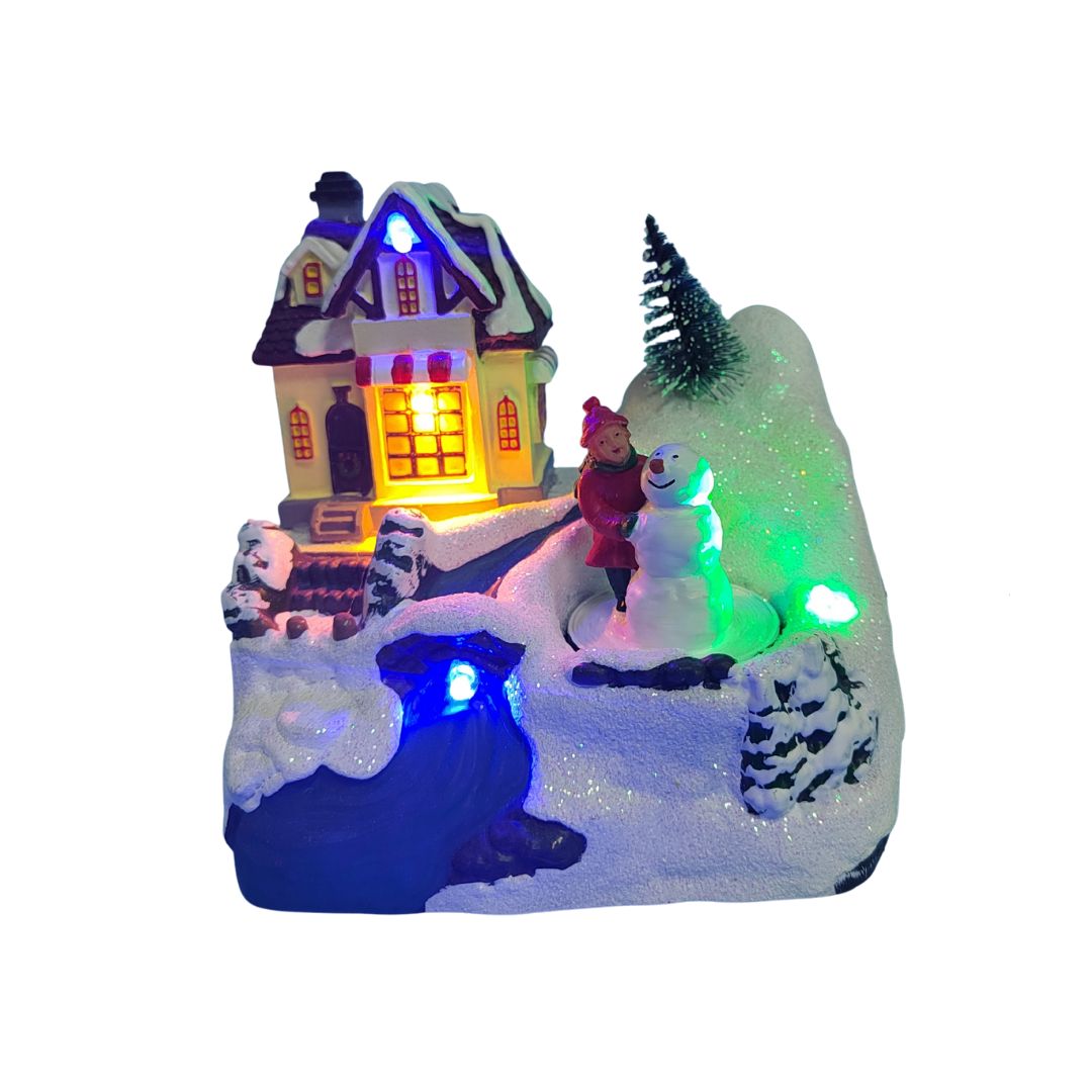 Villaggio natalizio con casa illuminata, bambina accanto a un pupazzo di neve e decorazioni innevate con luci LED colorate.