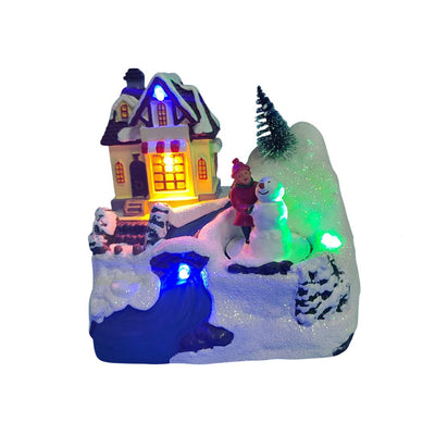 Villaggio natalizio con casa illuminata, bambina accanto a un pupazzo di neve e decorazioni innevate con luci LED colorate.
