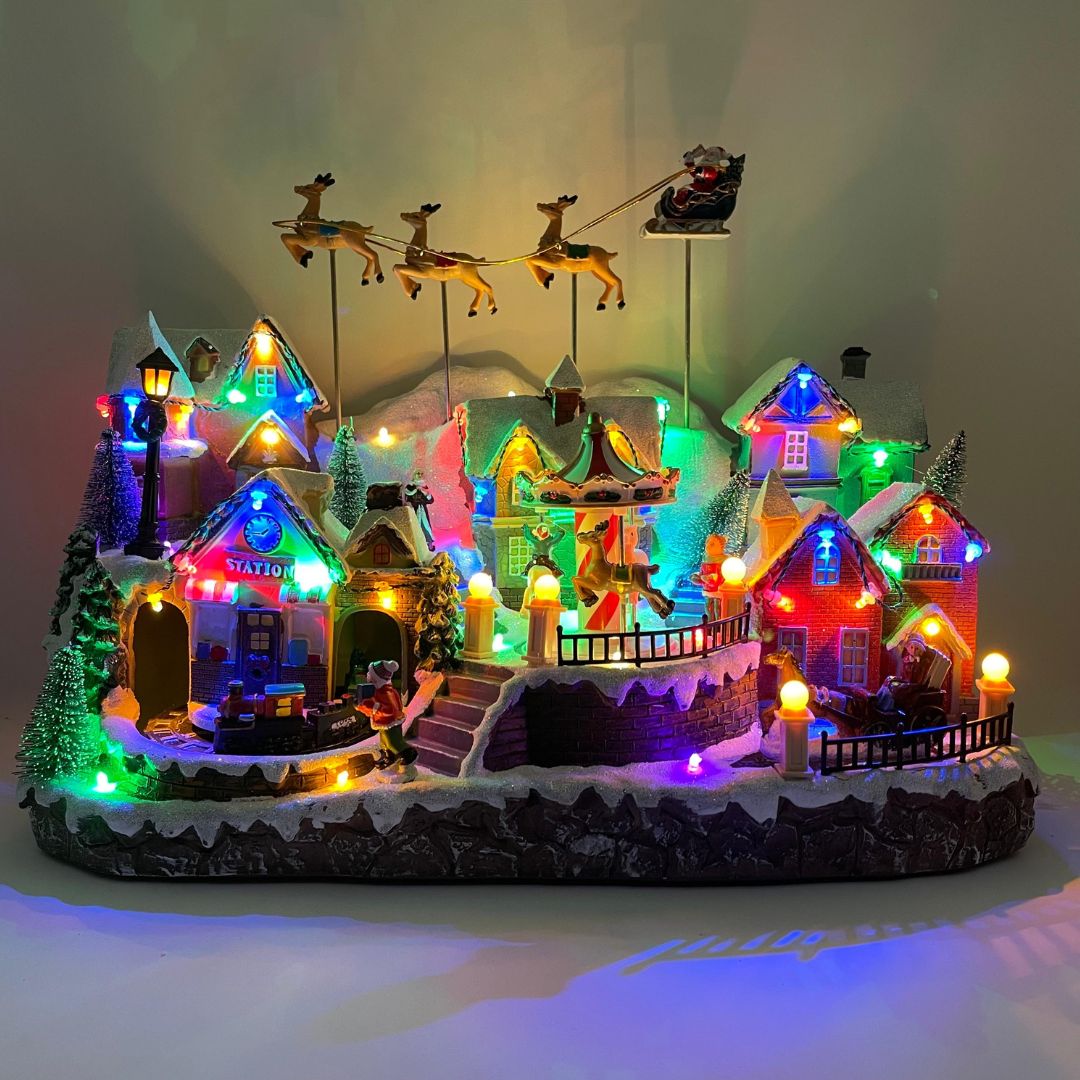 Villaggio natalizio luminoso con case innevate, giostra, trenino e slitta di Babbo Natale trainata da renne, illuminato da luci LED multicolore.