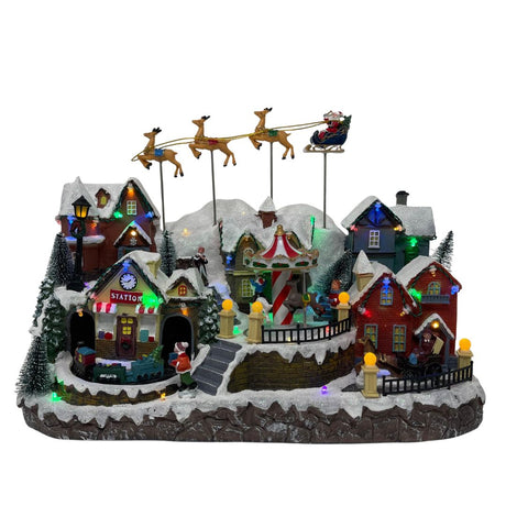 Villaggio natalizio luminoso con case innevate, giostra, trenino e slitta di Babbo Natale trainata da renne, illuminato da luci LED multicolore.