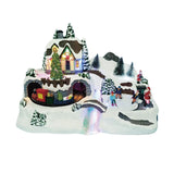 Villaggio natalizio in miniatura con casa illuminata, albero decorato, trenino che passa in galleria e figure di persone che pattinano sulla neve con Babbo Natale.