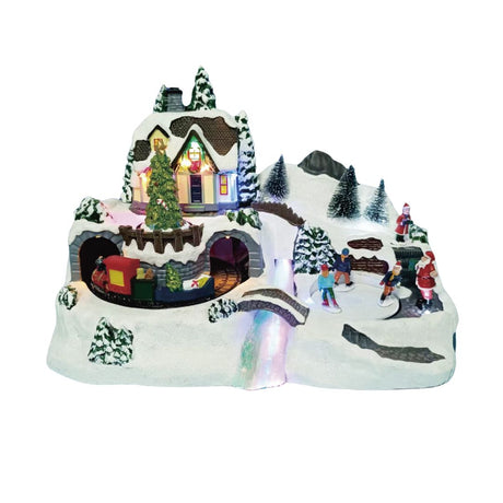 Villaggio natalizio in miniatura con casa illuminata, albero decorato, trenino che passa in galleria e figure di persone che pattinano sulla neve con Babbo Natale.